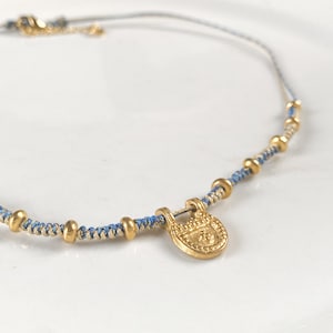 May include: A gold necklace with a blue and white cord and gold beads. The necklace has a gold pendant in the shape of a shield.