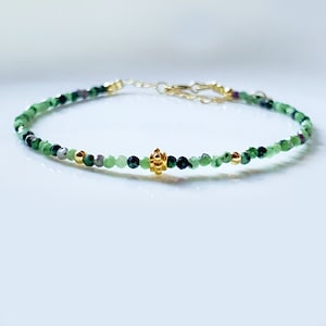以下が含まれることがあります： 緑色の宝石ビーズと金のアクセントでできた繊細なブレスレット。ブレスレットの中央には小さな金のチャームが付いています。