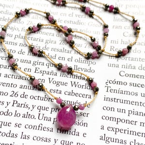 May include: A delicate necklace featuring a teardrop-shaped, faceted ruby pendant. The necklace is strung on a thin, beige cord, interspersed with small, pink and dark gray beads. The background is a printed page.
