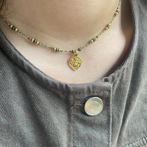 May include: A gold necklace with a small, ornate pendant in the shape of a leaf. The necklace is made of a thin, dark cord with small, black beads.