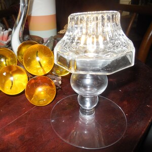 May include: A clear glass candle holder with a fluted top, a spherical stem, and a wide circular base. Beside it sits a cluster of translucent amber-colored glass grapes, featuring rounded spheres and a textured surface finish.