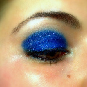 Op de afbeelding: Close-up van een oog met helderblauwe oogschaduw. De oogschaduw is aangebracht op het ooglid en heeft een glinsterend, glanzend effect. Het oog heeft donkere wimpers en een bruine iris. De wenkbrauw is donker en goed gedefinieerd.