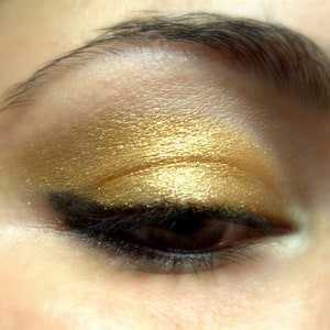 Könnte beinhalten: Nahaufnahme eines Frauenauges mit goldenem Lidschatten und schwarzem Eyeliner. Der Lidschatten ist schimmernd und hat einen goldenen Glanz.
