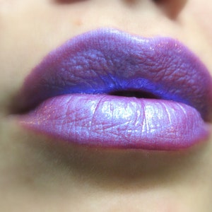 May include: Close-up of a person's lips wearing a shimmery purple lipstick with a blue undertone.