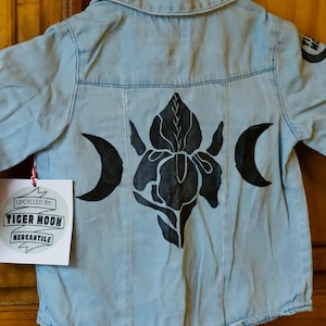 May include: Light blue denim shirt with long sleeves, featuring a black floral design and crescent moons on the back. A tag reads "Upcycled by Tiger Moon Mercantile."