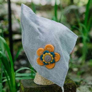 May include: A gray bonnet with a decorative felt flower in shades of orange, blue, and yellow, centered with a gray button. The bonnet is displayed outdoors, resting on a stone surface.