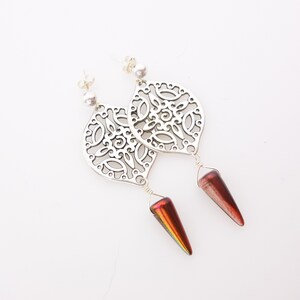 May include: A pair of silver-toned dangle earrings. Each earring features a filigree design in a leaf shape, with a small silver ball at the top. Below, a wire holds a dark red, cone-shaped bead. The beads have an iridescent sheen.