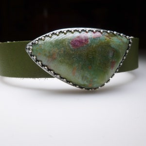 May include: A green and pink gemstone set in a silver bezel with a beaded edge. The gemstone is mounted on a green leather bracelet.
