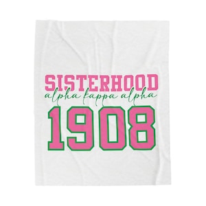 Op de afbeelding: Witte deken met de tekst "SISTERHOOD alpha kappa alpha 1908" in roze en groen. De deken is opgevouwen en heeft een textuur.