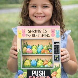 Puede incluir: Una máquina expendedora con temática de Pascua con el texto "SPRING IS THE BEST THING" y "PUSH EASTER". La máquina mide 28 cm de ancho y presenta huevos de Pascua decorados. El fondo es verde borroso.