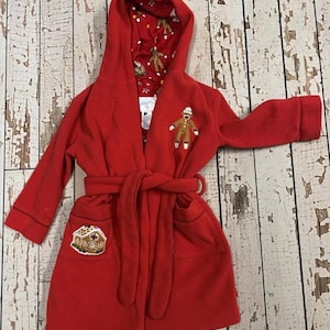 May include: A red children's robe with a gingerbread man and house applique. The robe has a hood with a patterned lining featuring gingerbread men and candy canes. It includes a matching belt and pockets.