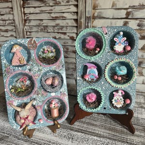 May include: Two blue and white cupcake tins with various Easter-themed decorations in each of the cups. The tins are on wooden stands. The decorations include birds, bunnies, gnomes, eggs, and flowers.
