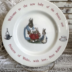 May include: A white ceramic plate with a red border featuring a depiction of three rabbits from the children's book "The Tale of Peter Rabbit". The plate has the text "Welcome to the new baby of the Prince and Princess of Wales, with love, Peter Rabbit."