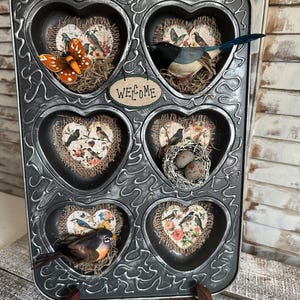 May include: A black metal heart-shaped muffin tin with six individual heart-shaped molds. Each mold is decorated with a different vintage-style paper heart featuring birds and flowers. The tin is decorated with a sign that says "WELCOME".