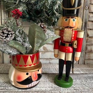 May include: A decorative Christmas-themed arrangement featuring a red and gold nutcracker soldier figurine and a matching planter. The planter is shaped like a nutcracker head. The background includes greenery and a string of lights.