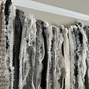 K&ouml;nnte beinhalten: Eine wei&szlig;e und graue Stoffgirlande mit verschiedenen Texturen, darunter Fransen, H&auml;kelarbeiten und Spitze. Die Girlande h&auml;ngt an einer wei&szlig;en Deckenleiste.