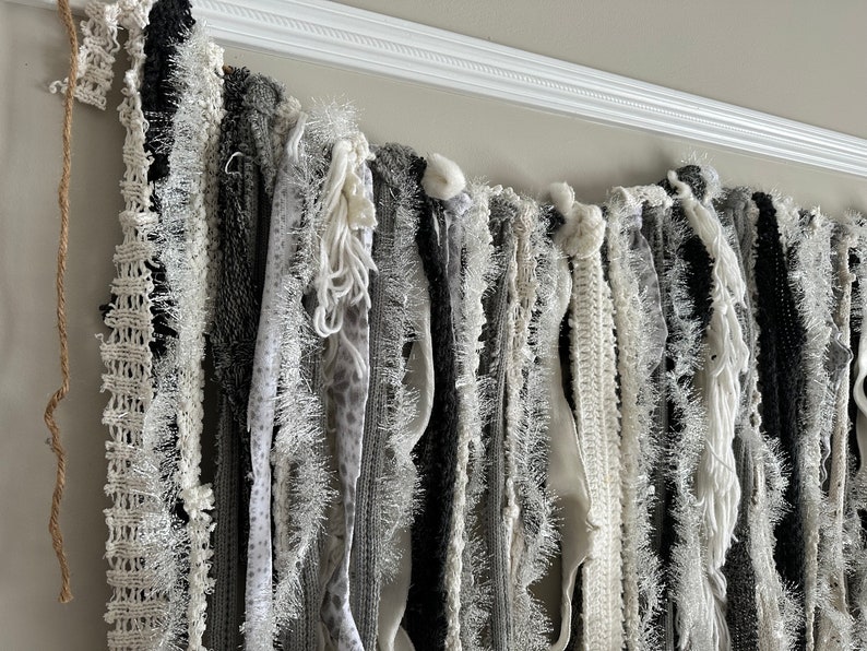 K&ouml;nnte beinhalten: Eine wei&szlig;e und graue Stoffgirlande mit verschiedenen Texturen, darunter Fransen, H&auml;kelarbeiten und Spitze. Die Girlande h&auml;ngt an einer wei&szlig;en Deckenleiste.