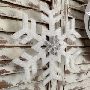 K&ouml;nnte beinhalten: Ein wei&szlig;er Schneeflocken-Anh&auml;nger mit einer silbernen Glitzer-Schneeflocke in der Mitte. Der Anh&auml;nger h&auml;ngt an einer Schnur vor einem wei&szlig;en Holz-Hintergrund.
