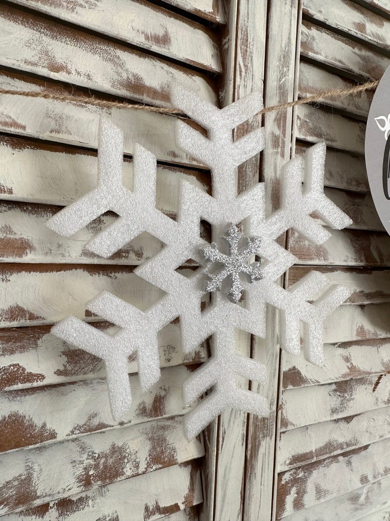 K&ouml;nnte beinhalten: Ein wei&szlig;er Schneeflocken-Anh&auml;nger mit einer silbernen Glitzer-Schneeflocke in der Mitte. Der Anh&auml;nger h&auml;ngt an einer Schnur vor einem wei&szlig;en Holz-Hintergrund.