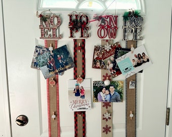 May include: A white door decorated with Christmas cards and wooden signs that read "Noel", "Hope", "Snow", and "Hope". The cards are held in place with burlap ribbon and wooden clothespins.