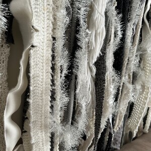 K&ouml;nnte beinhalten: Ein Wandbehang aus verschiedenen wei&szlig;en und grauen Stoffstreifen, darunter geh&auml;kelte und gefranste St&uuml;cke. Die Streifen sind vertikal angeordnet und erzeugen einen strukturierten, geschichteten Effekt.