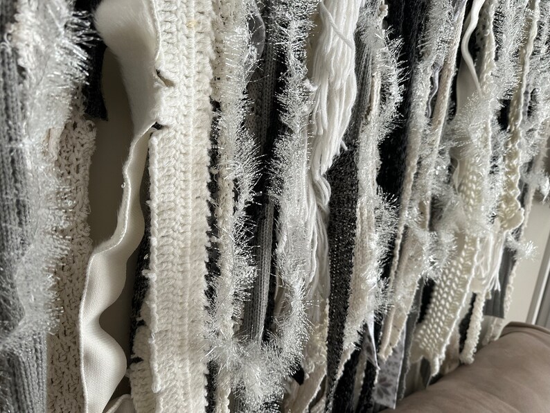 K&ouml;nnte beinhalten: Ein Wandbehang aus verschiedenen wei&szlig;en und grauen Stoffstreifen, darunter geh&auml;kelte und gefranste St&uuml;cke. Die Streifen sind vertikal angeordnet und erzeugen einen strukturierten, geschichteten Effekt.