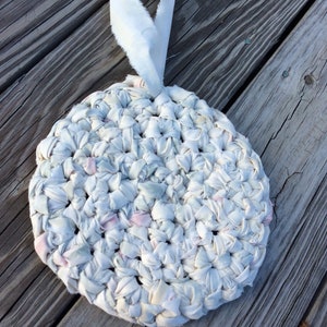 Puede incluir: Una alfombra redonda de crochet blanco con un lazo de tela blanco para colgar. La alfombra está hecha de tiras de tela tejidas en blanco, rosa y azul claro.