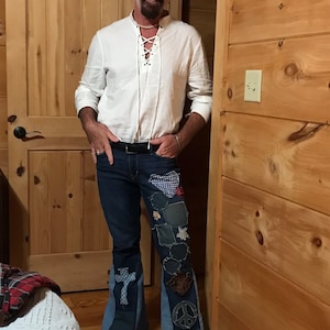 Op de afbeelding: Een man draagt een wit shirt met een veterkraag, een zwarte beanie met een witte rand en een blauwe jeans met patchwork details. De jeans heeft een wijde pijp en verschillende stoffen patches, waaronder een vredesteken, een kruis en een ster.