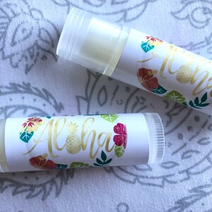 May include: Two white lip balm tubes with a tropical floral design and the word "Aloha" in gold lettering. The design includes green leaves, pink flowers, and a pineapple.