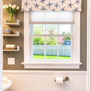 May include: A bathroom with a window featuring a white frame and a view of a green yard. The window is adorned with a valance patterned with blue starfish on a white background. White shelves hold a vase of tulips, a decorative jar, and a rolled towel.