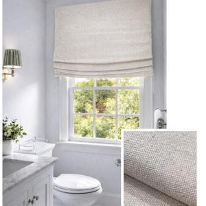May include: A neutral-toned Roman shade made of textured fabric is installed above a window. The window is divided into multiple panes, offering a view of lush greenery. The bathroom features a white vanity, toilet, and wall-mounted sconce.