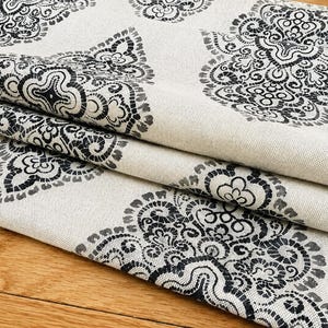 May include: Cream-colored fabric with a repeating black floral pattern. The fabric is folded, revealing the texture and design. The pattern features intricate, symmetrical floral motifs, creating a decorative textile.