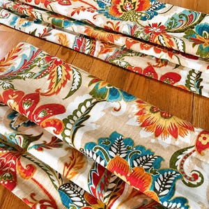 May include: Rolled fabric with a vibrant floral pattern in red, orange, blue, and green on a beige background. The fabric is arranged on a wooden surface, showcasing the detailed design and color variations.