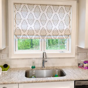 May include: A kitchen window with a white frame and a patterned Roman shade. The shade features a repeating diamond design in beige and white. A stainless steel sink and faucet are visible below the window, with a granite countertop.