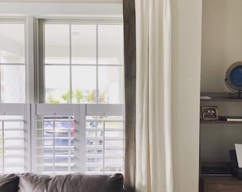 May include: White curtains with brown trim hang from a wooden rod in front of a window with white shutters. A brown leather couch sits in front of the window.
