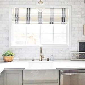 May include: A white kitchen with white subway tile backsplash, a white farmhouse sink, and a stainless steel dishwasher. The window has a white roman shade with black stripes. There are two framed prints on the wall above the sink.