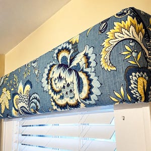 May include: A blue floral valance with large, stylized flowers in shades of yellow, white, and brown. The valance is mounted above white blinds, creating a decorative window treatment. The fabric has a linen-like texture.