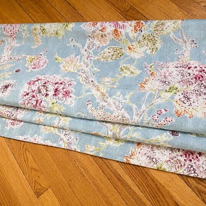May include: A folded fabric with a light blue background and a floral pattern. The design features large, detailed flowers in shades of pink, purple, and orange, with green and white foliage. The fabric is lying on a wooden surface.