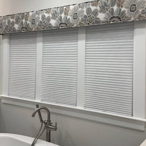 May include: A bathroom scene featuring a white bathtub and window blinds. The window has three sections with pleated blinds. Above the window is a floral valance in gray, white, and brown. A silver faucet is visible.