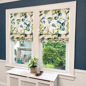 May include: Two windows with white frames and blue walls. Each window has a Roman shade with a floral pattern in blue, green, and cream. A small potted plant sits on a white radiator cover below the windows.