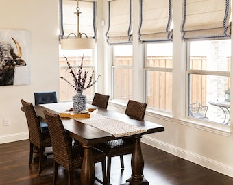 May include: A dining room with a dark wood table and six chairs. The chairs are a mix of woven and upholstered styles. The room has three windows with cream-colored shades trimmed with blue. A chandelier hangs above the table, and a painting of a bull is on the wall.