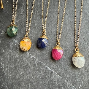 May include: A collection of gold-toned necklaces, each featuring a unique gemstone pendant. The pendants are in various colors, including green, yellow, blue, pink, and white. The gemstones are faceted and set in gold-colored caps, suspended from delicate gold chains.