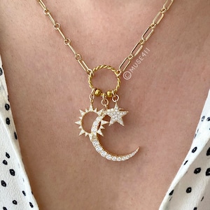Op de afbeelding: Een gouden ketting met een bedel in de vorm van een halve maan en een zon met een ster erboven. De bedels zijn versierd met glinsterende, heldere stenen.