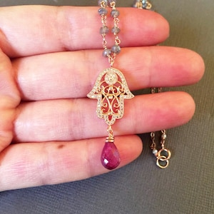 Puede incluir: Un collar de oro con un dije de mano de Hamsa y un colgante de piedra preciosa de rubí. El collar está hecho con una cadena de cuentas grises y una cadena de oro.