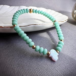 Peut inclure: Un bracelet délicat composé de perles de verre vert clair et d'un charme hamsa doré avec une pierre d'opale blanche.