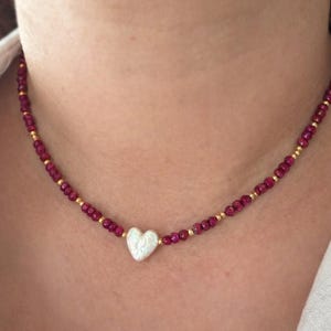 Może przedstawiać: Delikatny naszyjnik z białą zawieszką w kształcie serca. Naszyjnik jest nawleczony z małych, okrągłych, ciemnoczerwonych koralików i przeplatany maleńkimi, złotymi koralikami. Naszyjnik jest noszony na szyi.