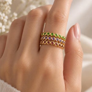 Könnte beinhalten: Ein Stapel aus drei Goldringen, die jeweils mit einer Reihe kleiner, runder, in Zargenfassung gesetzter Edelsteine besetzt sind. Der oberste Ring hat grüne Steine, der mittlere violette und der untere rote Steine in einem Goldband.