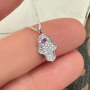 May include: A silver necklace with a hamsa hand pendant. The pendant is covered in small clear stones and has a pink stone in the centre.