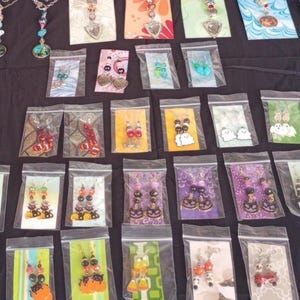 May include: A variety of Halloween-themed earrings displayed in individual clear plastic bags. The earrings feature different designs, including ghosts, pumpkins, candy corn, and black cats. The earrings are made with colorful beads and charms.