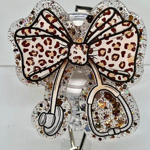 May include: A clear glitter badge reel with a brown and white leopard print bow and a stethoscope. The reel has a silver clip and a silver ring.